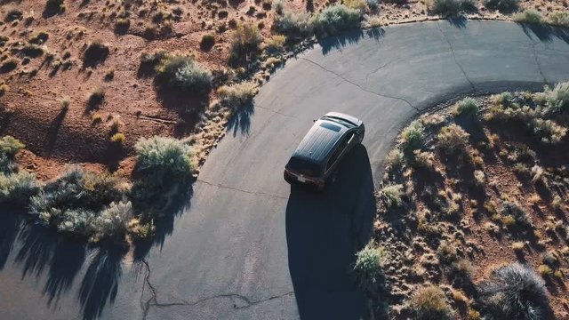 Top view drone follows silver minivan car taking turns on small narrow asphalt desert ring road on a sunny summer day.