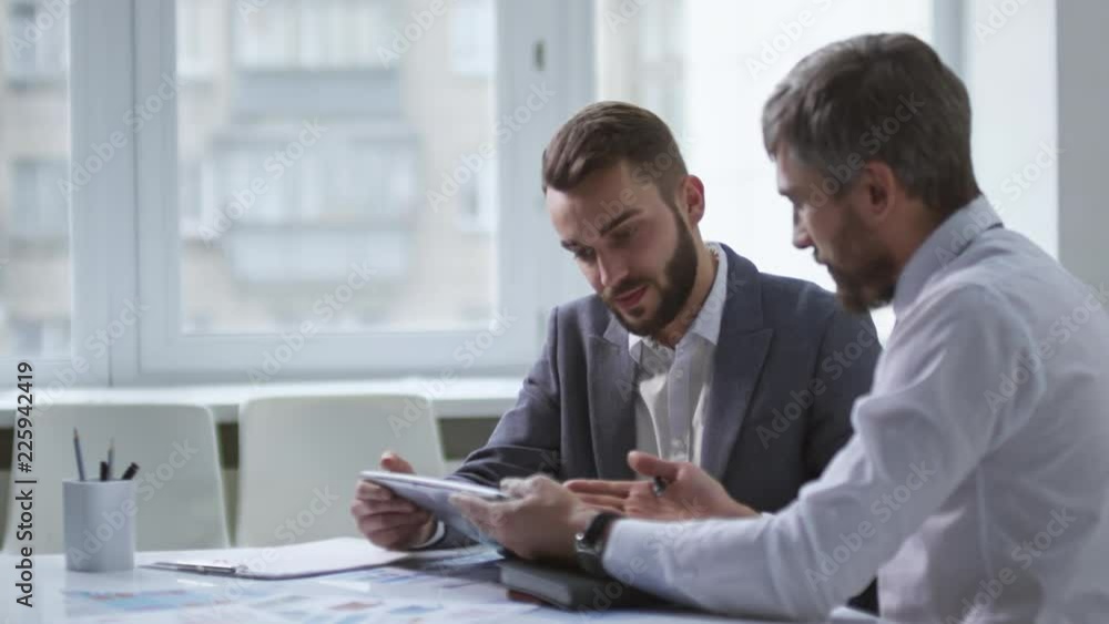 Poster side view of two male colleagues discussing work details using tablet computer when sitting at desk 