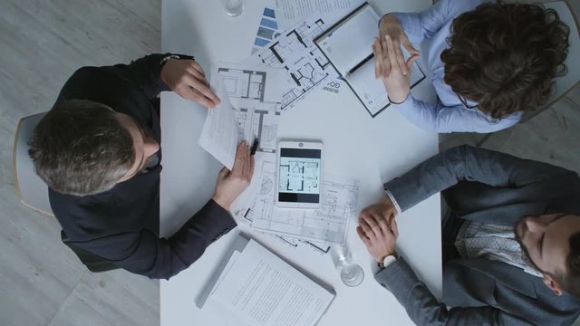 Top view of male real estate agent showing contract and technical drawings to clients when sitting at desk with documents on it, zoom in shot