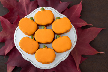 Naklejka na ściany i meble White platter with small orange frosted pumpkin cakes on brown background, with red fall leaves 