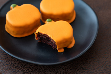 Black plate with small orange frosted pumpkin cakes, one with bite taken out, on brown background 
