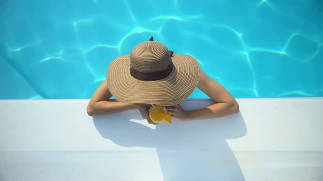 Young woman in elegant hat swimming in pool and enjoying delicious cocktail