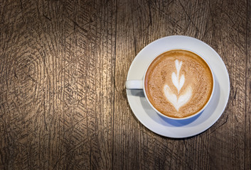 A white cup of cappuccino with a leaf shape on the top over a wooden background.