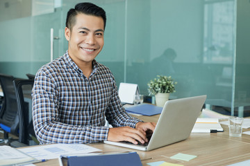 Smiling handsome developer working on laptop at his workplace in office