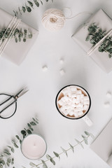 A mug with hot chocolate and marshmallow among gift boxes and eucalyptus on a white table. The concept of wintertime and wrapping gifts. Flat lay, top view, minimalist style.