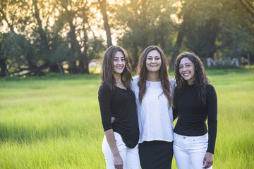 Horizontal Outdoor Portrait of three beautiful Hispanic young women standing together outdoors. Smiling and looking at a camera