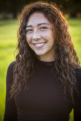 A beautiful Hispanic teen girl outdoor portrait. Cute and smiling woman with brown hair looking at the camera standing in a scenic field outdoors