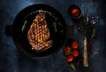 Medium rare grilled Steak Ribeye in a cast-iron pan, a fork for meat, herbs and spices on a dark concrete background