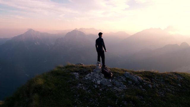 Aerial - Flying around young man standing victoriously on top of the mountain at sunset