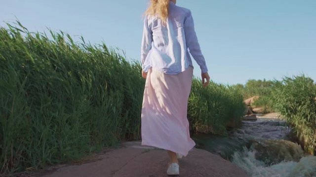 A young girl, blonde in a pink dress and blue shirt, is walking along the rocky shore of the river. The wind develops long, curly hair.