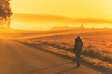 Fototapeta na wymiar Young woman in the winter jacket with a backpack on the road against the background of the sunrise autumn field. Travel and tourism concept