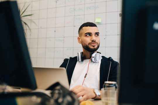Young male computer hacker sitting with laptop while looking away at creative office
