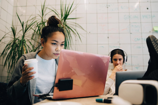 Confident young businesswoman holding coffee while using laptop by female colleague at creative office