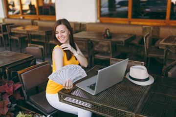 Woman in outdoors street coffee shop cafe sitting with modern laptop pc computer, hold in hand bunch of dollars banknotes, cash money. Mobile Office restaurant in free time. Freelance business concept