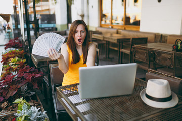 Woman in outdoors street coffee shop cafe sitting with modern laptop pc computer, hold in hand bunch of dollars banknotes, cash money. Mobile Office restaurant in free time. Freelance business concept