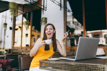 Happy girl in outdoors street coffee shop cafe sitting at table with laptop pc computer, texting message on mobile phone, drink cup tea in restaurant during free time. Mobile office freelance concept.