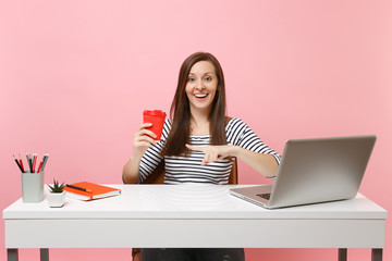 Young smiling woman pointing index finger on cup of coffee or tea working on project sit at office with pc laptop isolated on pastel pink background. Achievement business career concept. Copy space.