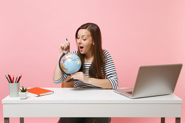 Curious woman pointing on globe with pencil, planning vacation while sit and work at white desk with contemporary pc laptop isolated on pastel pink background. Achievement business career. Copy space.