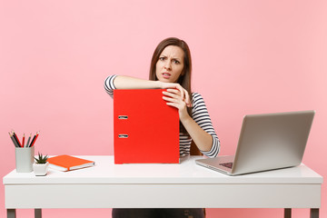 Puzzled woman leaning on red folder with paper documents and working on project while sitting at...