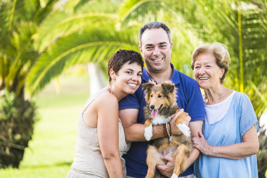 Family Group Of Caucasian Happy People With Different Ages From 30 To 80 Years Old And Nice Funny Shetland Puppy Dog With Them. Love And Friendship Concept For Cheerful People In Outdoor Activity