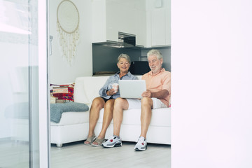 senior adult caucasian people couple sit down on the sofa at home use technology and internet with a laptop together. smile and relax together in an elegant white bright home. modern mature computer