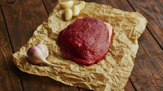Aromatic fresh garlic and piece of uncooked meat lying on parchment paper on scratched wooden tabletop