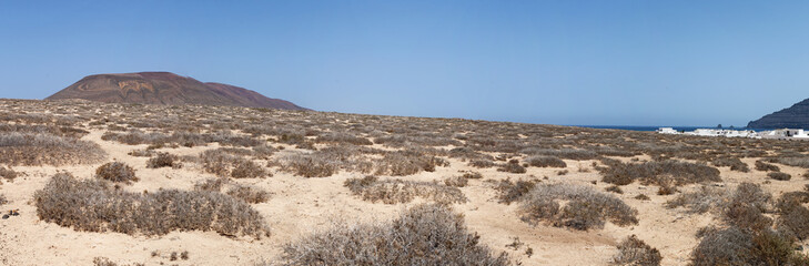 Fototapeta na wymiar Lanzarote, Isole Canarie: strada sterrata, cespugli e paesaggio desertico con la Montagna Pedro Barba, il vulcano dell'isola La Graciosa