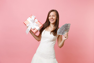 Portrait of laughing bride woman in white wedding dress hold bundle lots of dollars, cash money, red box with gift, present isolated on pink pastel background. Wedding celebration concept. Copy space.