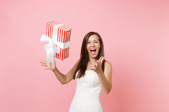 Funny joyful bride woman in elegant white wedding dress screaming throwing up red box with gift present isolated on pink pastel background. Wedding celebration concept. Copy space for advertisement.
