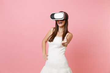 Portrait of joyful bride woman in white wedding dress, headset of virtual reality showing thumb up isolated on pastel pink background. Organization of wedding concept. Copy space for advertisement.