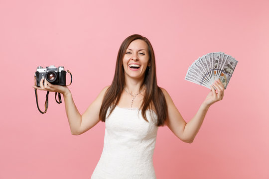 Laughing bride woman in wedding dress holding retro vintage photo camera, bundle lots of dollars, cash money choosing staff, photographer isolated on pink background. Organization, Wedding to do list.