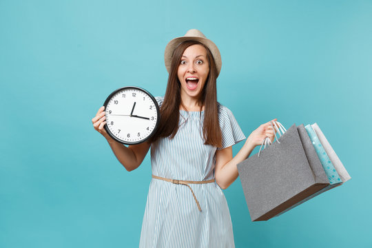 Portrait beautiful young caucasian woman in summer dress, straw hat holding packages bags with purchases after shopping, round clock isolated on blue pastel background. Copy space for advertisement.