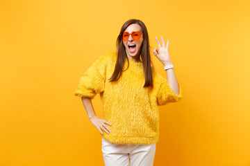 Portrait of joyful young woman in fur sweater, white pants and heart orange glasses showing OK sign isolated on bright yellow background. People sincere emotions, lifestyle concept. Advertising area.