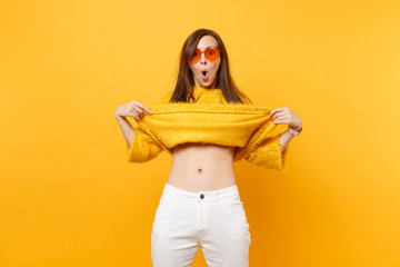 Portrait of shocked funny young woman in heart orange glasses taking off fur sweater, showing belly...
