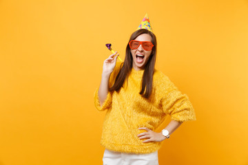 Excited happy young woman in orange funny eyeglasses birthday party hat with playing pipe...