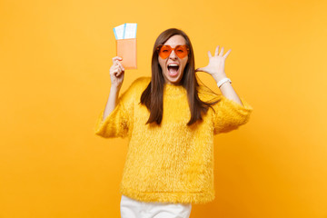 Excited happy young woman in orange heart glasses screaming spreading hands, holding passport boarding pass tickets isolated on yellow background. People sincere emotions, lifestyle. Advertising area.