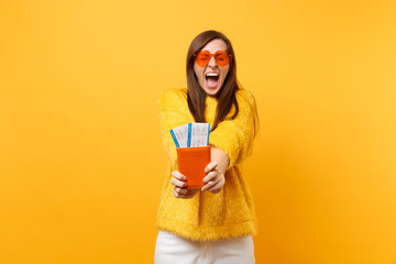Crazy pretty young woman with closed eyes in orange heart glasses screaming, holding passport boarding pass tickets isolated on yellow background. People sincere emotions, lifestyle. Advertising area. - obrazy, fototapety, plakaty