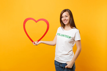 Portrait of woman in white t-shirt with written inscription green title volunteer hold big red wooden heart isolated on yellow background. Voluntary free assistance help, charity grace work concept.