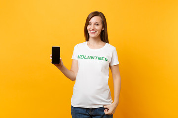 Woman in white t-shirt written inscription green title volunteer hold mobile phone with blank empty screen isolated on yellow background. Voluntary free assistance help, charity grace work concept.