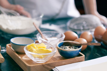 Close-up view of bowls with flour, butter and eggs for cookie dough prepared on kitchen counter, anonymous woman standing on blurred background