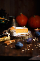 Piece of pumpkin cheesecake with powdered sugar, plums, pumpkins, table lamp on a dark wooden background