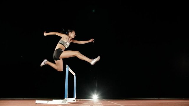 Runner heading forward. Young female athlete blasting off on short track of stadium and jumping over barriers, training before olympics 4k
