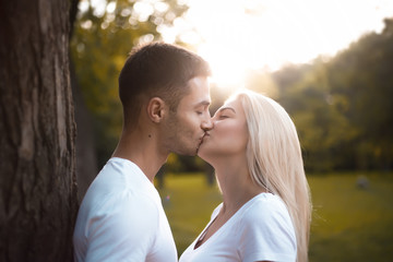 Happy young cute loving couple standing near tree in park kissing with beautiful sundown light.