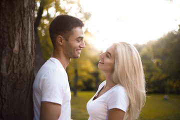 Young cute cheerful loving couple standing near tree in park looking at each other with beautiful sundown light.