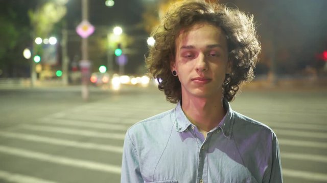  Exciting view of a young man with long blond hair walking along a zebra crossing and smiling cheerfully in a city street at night in autumn  