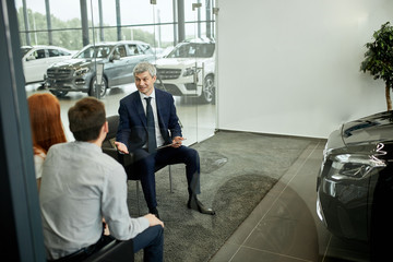 Caucasian couple is buying new car, mature seller in a formal wear is presenting contract to sign,...