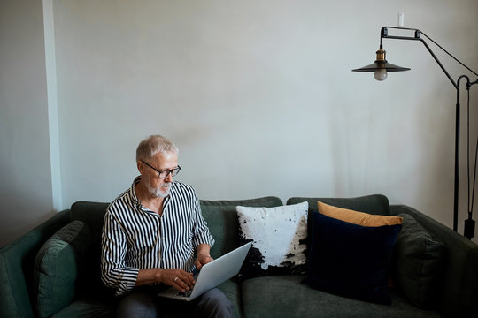 Trendy Mature Bearded Man Working From Home With Laptop. Sitting At Desk Near Window