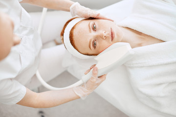 Cosmetology RF lifting equipment. Anti-age and anti-wrinkle apatherapy. Top view of beautiful woman in white bathrobe getting facial treatment and looking at camera at Cosmetology Salon.