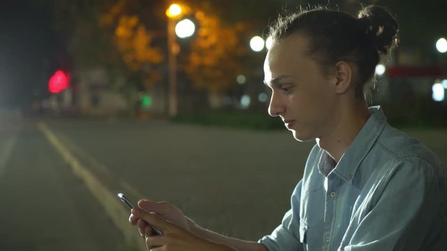  Profile of an elegant young man with a bun haircut browsing the net cheery on his smartphone and seeking info in a city street deep at night in autumn