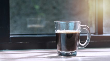 Hot Black coffee in glass beside window of the morning on wooden background.